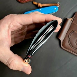 Puede incluir: Una cartera de cuero negro con detalles de costura roja y un bot&oacute;n de metal dorado. La cartera est&aacute; abierta y muestra varias tarjetas en el interior.