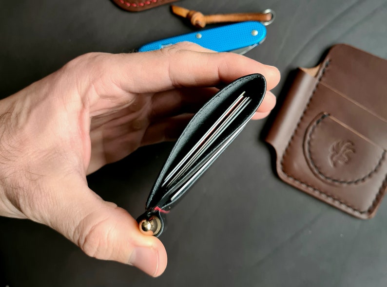 Puede incluir: Una cartera de cuero negro con detalles de costura roja y un bot&oacute;n de metal dorado. La cartera est&aacute; abierta y muestra varias tarjetas en el interior.