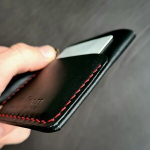 Puede incluir: Cartera de cuero negro con costuras rojas y una ranura para tarjeta. La cartera tiene la palabra "Icon" grabada en relieve en la parte delantera.