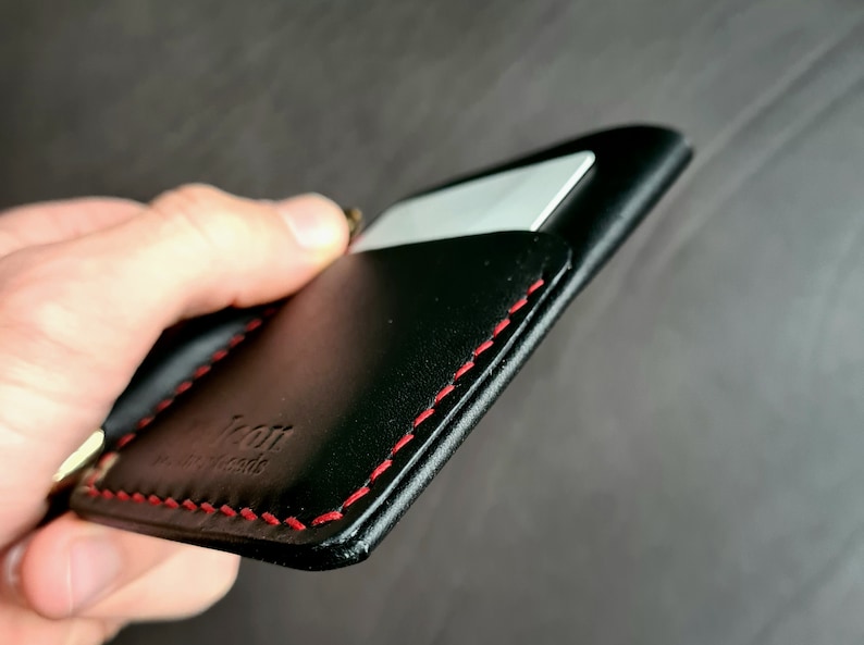 Puede incluir: Cartera de cuero negro con costuras rojas y una ranura para tarjeta. La cartera tiene la palabra "Icon" grabada en relieve en la parte delantera.