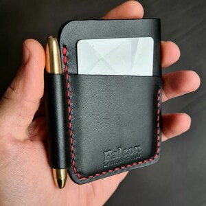 Puede incluir: Cartera de cuero negro con detalles de costura roja y un portaplumas. La cartera tiene una ranura para tarjetas y el texto "Falcon Leather Goods" est&aacute; estampado en la parte delantera.