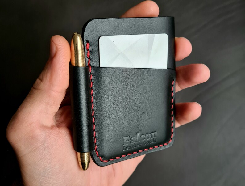 Puede incluir: Cartera de cuero negro con detalles de costura roja y un portaplumas. La cartera tiene una ranura para tarjetas y el texto "Falcon Leather Goods" est&aacute; estampado en la parte delantera.