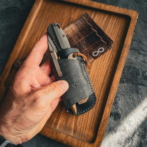 May include: A grey pocket knife and a brass pen are held in a black leather sheath with a decorative emblem. A brown and black plaid pouch with an infinity symbol is also visible. The items are displayed on a wooden tray.