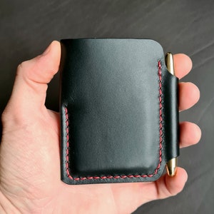 Puede incluir: Cartera de cuero negro con una costura roja y un portaplumas. La cartera se sostiene en una mano.