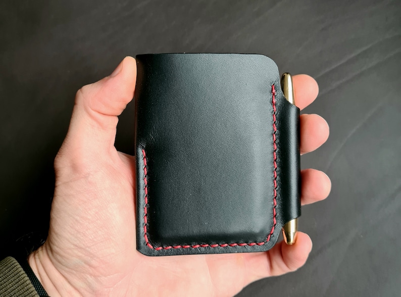 Puede incluir: Cartera de cuero negro con una costura roja y un portaplumas. La cartera se sostiene en una mano.