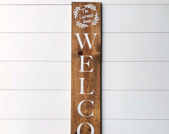 May include: A wooden welcome sign with the text "WELCOME" in white letters. The sign also features a wreath with the text "The Laurence home" in the center.