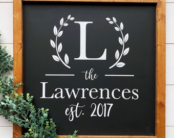 May include: A framed wooden sign with a black background features the white text "L" inside a laurel wreath, "the Lawrences est. 2017". The frame is a light brown color. A green leafy garland is in the lower left corner.