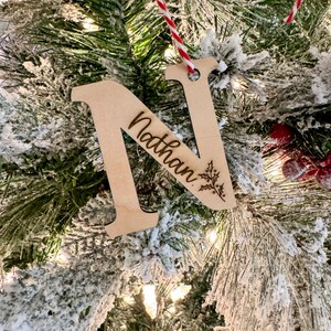 May include: A wooden Christmas ornament shaped like the letter N with the name "Nathan" engraved on it. The ornament is hanging on a snowy Christmas tree.