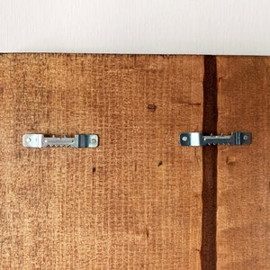May include: The back of a wooden sign with two metal sawtooth hangers attached. The wood is a light brown color.
