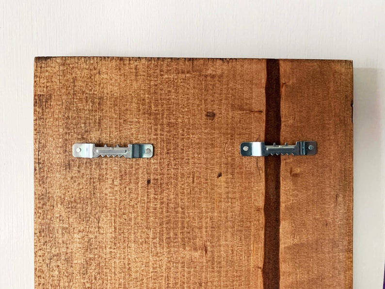 May include: The back of a wooden sign with two metal sawtooth hangers attached. The wood is a light brown color.