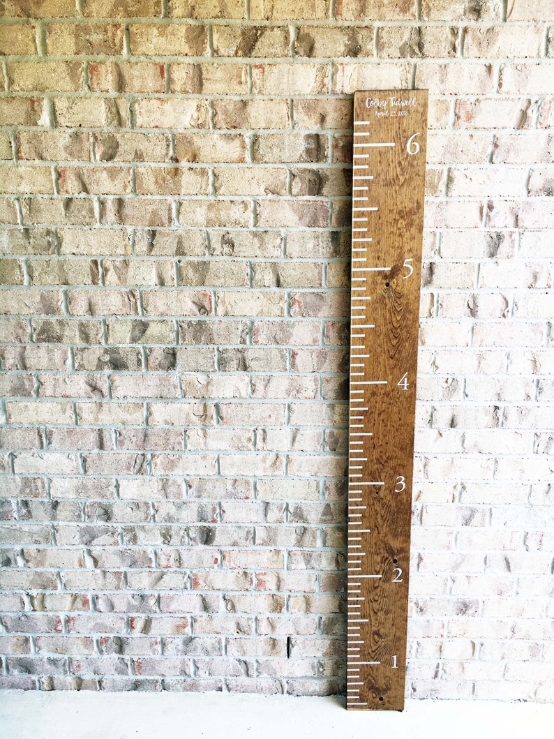 May include: A wooden growth chart with white markings and numbers. The chart is stained a dark brown color and has the name "Colby Tisdell" and the date "April 21, 2018" written on it.
