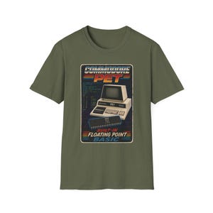 May include: An olive green t-shirt featuring a graphic print of a vintage Commodore PET computer. The design includes the text COMMODORE PET, BUILT-IN FLOATING POINT BASIC, and a detailed illustration of the computer hardware on a dark background.