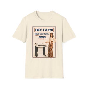 May include: A cream-colored short-sleeve t-shirt featuring a graphic print of a vintage DEC LA 120 printer and a person pointing at it. The text reads DEC LA 120, Work from Home, digital, and DECWRITER III. The print is framed in a thin red border.