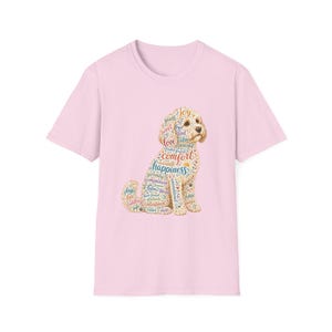 Puede incluir: Una camiseta de manga corta de color rosa claro con un gráfico de un perro sentado compuesto por textos coloridos y en espiral. Las palabras incluyen joy, sweet, love, comfort, happiness, serenity, companion y otros términos positivos.