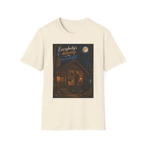 Könnte beinhalten: Ein cremefarbenes Kurzarm-T-Shirt mit einem rechteckigen Grafikdruck einer Holzhütte bei Nacht. Die Szene zeigt zwei Figuren, die in der beleuchteten Hütte tanzen, mit dem Text "Everybody's dancing in the moonlight" darüber.