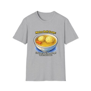 May include: A heather gray t-shirt featuring a graphic of a bowl of matzo ball soup with two matzo balls and a carrot slice. The text above reads Matzo Ball Soup: and the text below reads Chicken and Dumplings With Attitude in black letters.