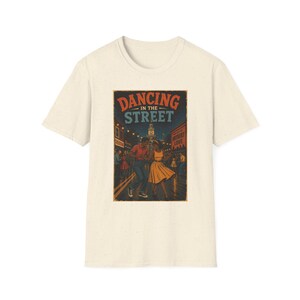 May include: A cream-colored short-sleeve t-shirt featuring a graphic print of a couple dancing on a city street at night. The text DANCING IN THE STREET is printed in orange and blue above the illustration, which includes a clock tower.