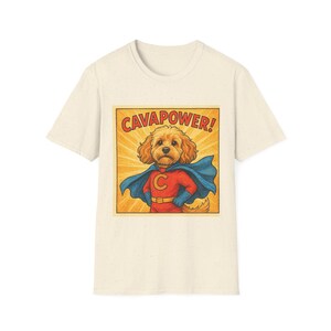 Op de afbeelding: Een crèmekleurig T-shirt met korte mouwen en een grafische print van een goudbruine hond verkleed als superheld met een blauwe cape en een rood pak. De tekst CAVAPOWER! staat in het rood boven de hond op een gele achtergrond.