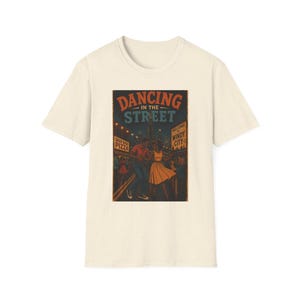 May include: A cream-colored short-sleeve t-shirt featuring a graphic print of a couple dancing on a city street. The text reads DANCING IN THE STREET, DEEP DISH PIZZA, and WELCOME TO WINDY CITY. The illustration uses a vintage color palette.