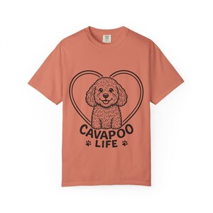 Op de afbeelding: Een koraalkleurig T-shirt met korte mouwen en een zwarte afbeelding van een hond met krullend haar, gecentreerd in een hartvormige omtrek. Onder de hond staat de tekst CAVAPOO LIFE, vergezeld door twee kleine pootafdrukken naast het woord LIFE.