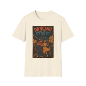 May include: A cream-colored short-sleeve t-shirt featuring a graphic print of a couple dancing on a city street at night. The text reads DANCING IN THE STREET, DEEP DISH PIZZA, and WELCOME TO WINDY CITY. The illustration uses warm tones against a dark background.