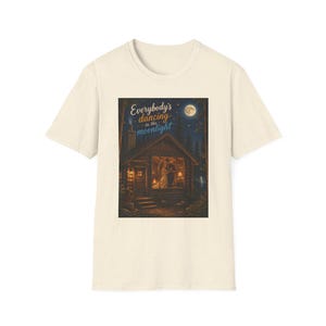 Könnte beinhalten: Ein cremefarbenes Kurzarm-T-Shirt mit einem Grafikdruck einer Blockhütte bei Nacht unter einem Vollmond. Der Text "Everybody's dancing in the moonlight" ist über der Hütte aufgedruckt, in deren Fenster ein tanzendes Paar zu sehen ist.
