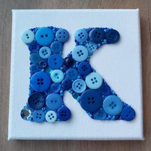 Puede incluir: Un lienzo blanco con una letra K azul hecha de botones. Los botones son de varios tonos de azul y algunos tienen un centro blanco.