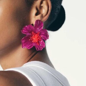 Puede incluir: Un pendiente grande en forma de flor de color magenta vibrante con pétalos translúcidos superpuestos y un centro texturizado de color naranja brillante. El pendiente está sujeto al lóbulo, mostrando su diseño audaz.