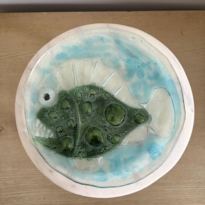 May include: A circular glass plate with a green fish design. The fish has a textured body with dark green spots and a wide grin. The plate's rim is white, with a light blue water-like design. The plate is likely for decorative purposes.