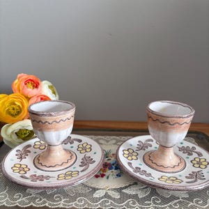 May include: Two ceramic egg cups with matching saucers. The egg cups are white with a brown band and sit on a pedestal base. The saucers are white with a floral design in yellow, brown, and green. The set is on a glass tray.