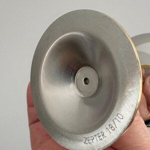 May include: Close-up of a stainless steel object with a brushed finish. The object has a conical shape with a small hole at the center. The text "ZEPTER 18/10" is etched on the surface. The rim has a gold-colored band.
