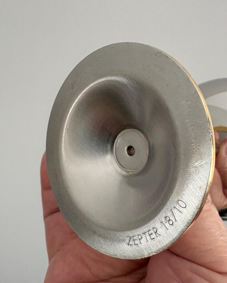 May include: Close-up of a stainless steel object with a brushed finish. The object has a conical shape with a small hole at the center. The text "ZEPTER 18/10" is etched on the surface. The rim has a gold-colored band.