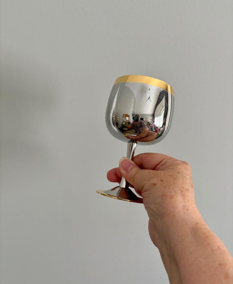 May include: A shiny, silver wine glass with a gold rim and base. The glass is held up, reflecting the surrounding environment. The stem is slender and elegant, adding to the overall sophisticated design of the glassware.