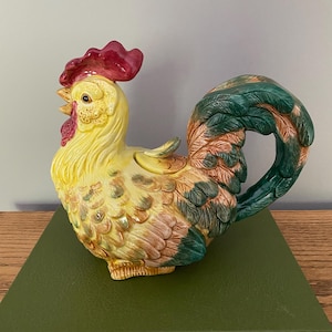 May include: A ceramic rooster teapot with a yellow body, red comb, green and brown tail feathers, and a lid. The teapot is sitting on a green book.