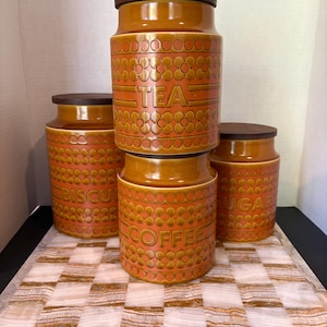 May include: A set of four cylindrical ceramic kitchen canisters in a warm orange hue, each featuring a dark brown wooden lid and a repeating embossed floral pattern. The canisters are labeled with the words TEA, COFFEE, SUGAR, and BISCUITS.