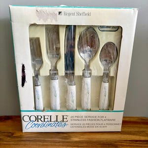 May include: A boxed set of Corelle Coordinates 20-piece stainless steel flatware. The set includes a knife, fork, spoon, and teaspoon, each with a white handle adorned with a blue floral design. The box reads "Regent Sheffield".