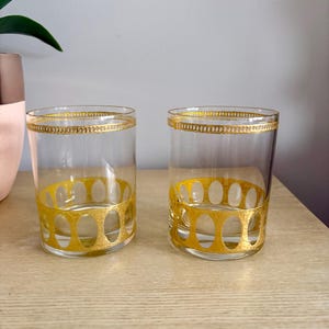 May include: Two clear drinking glasses with gold-colored decorative bands. The glasses have a gold band around the top rim and a gold oval pattern around the base. The glasses are cylindrical and sit on a light-colored wooden surface.