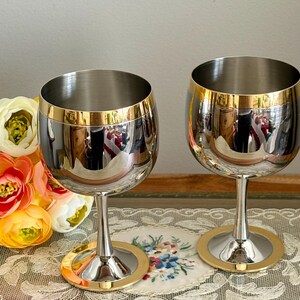 May include: Two stainless steel wine glasses with gold rims and bases. The glasses have a reflective surface, and the stems are slender. The background includes a floral arrangement and a decorative lace cloth.