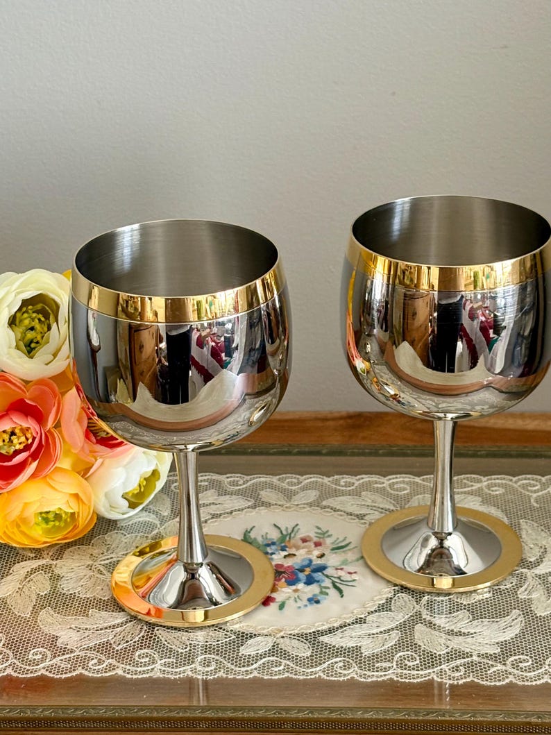 May include: Two stainless steel wine glasses with gold rims and bases. The glasses have a reflective surface, and the stems are slender. The background includes a floral arrangement and a decorative lace cloth.