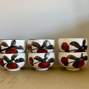May include: Six small white ceramic bowls with a speckled glaze. Each bowl features a black and red design of a branch with red fruit. The bowls are stacked in pairs.