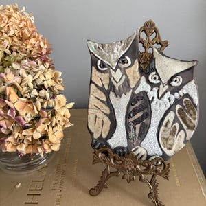 May include: A ceramic owl plaque featuring two owls with brown, beige, and white details. The plaque is displayed on a small, ornate brass easel. The owls have detailed feather patterns and are set against a cracked white background.