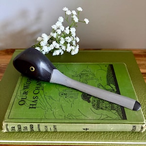 May include: A wooden letter opener with a dark grey head and a light grey handle, resting on a stack of green books. The letter opener has a gold-coloured accent. A sprig of white flowers is placed near the letter opener.