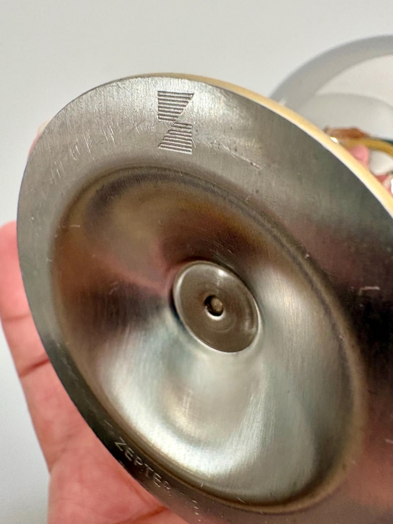May include: Close-up of a stainless steel base of a Zepter product. The base is circular with a central indentation and a visible logo. The word "ZEPTER" is etched on the side of the base.