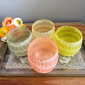 May include: Four colorful glass bowls with a delicate white web-like design. The bowls are in shades of green, yellow, peach, and tan. They sit on a decorative tray with a lace doily. A bouquet of flowers is in the background.