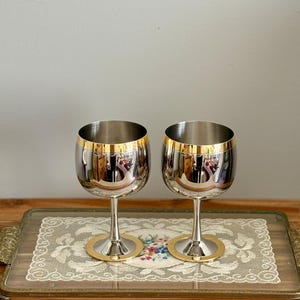May include: Two stainless steel wine glasses with gold rims and bases. The glasses are on a decorative tray with a floral design and lace trim. The tray has ornate gold handles and is set on a wooden surface.