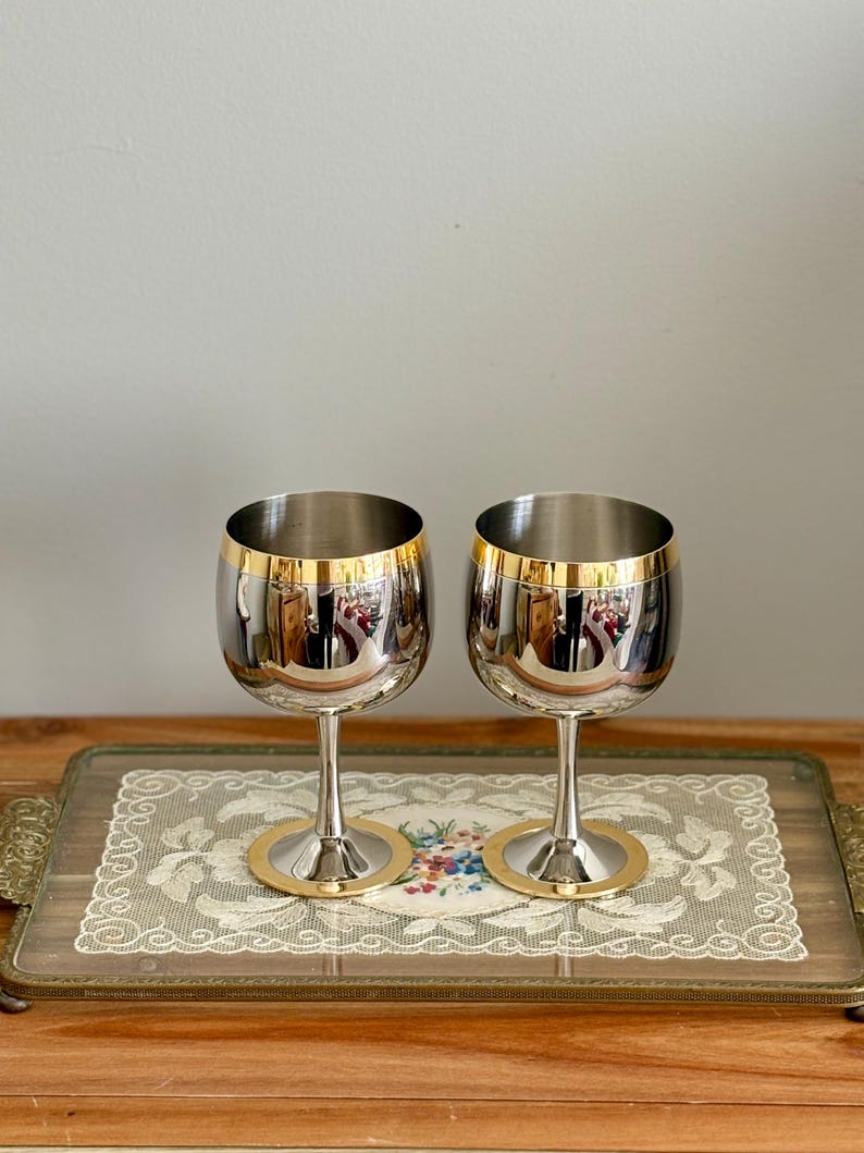 May include: Two stainless steel wine glasses with gold rims and bases. The glasses are on a decorative tray with a floral design and lace trim. The tray has ornate gold handles and is set on a wooden surface.