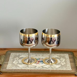 May include: Two stainless steel wine glasses with gold rims and bases. The glasses are displayed on a decorative tray with a floral design. The glasses reflect the surrounding environment.