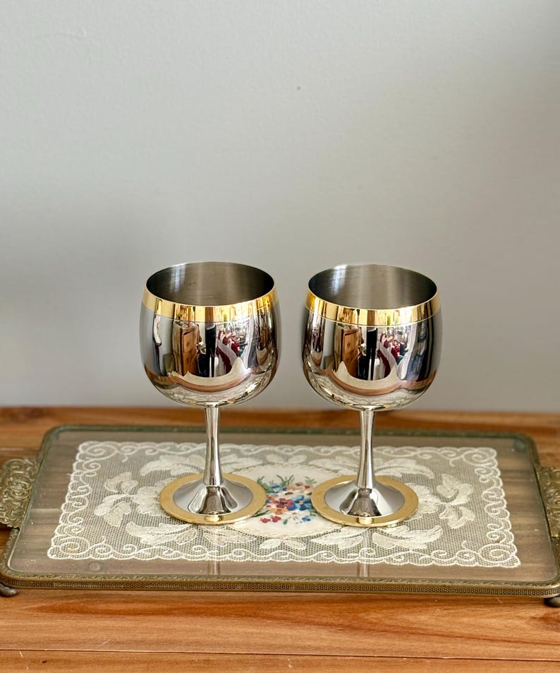 May include: Two stainless steel wine glasses with gold rims and bases. The glasses are displayed on a decorative tray with a floral design. The glasses reflect the surrounding environment.