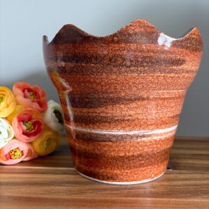 May include: A ceramic planter with a wavy rim, featuring horizontal bands of brown and orange hues. A white stripe accents the middle. The textured surface gives a rustic appearance. The planter is empty and sits on a wooden surface.