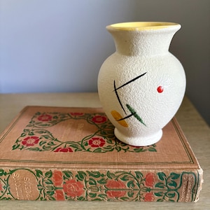 May include: A small, off-white ceramic vase with a textured surface, yellow interior, and abstract black, green, and yellow design. A red dot is on the side. The vase sits on a vintage book with floral illustrations and the text "Milton's Poems".